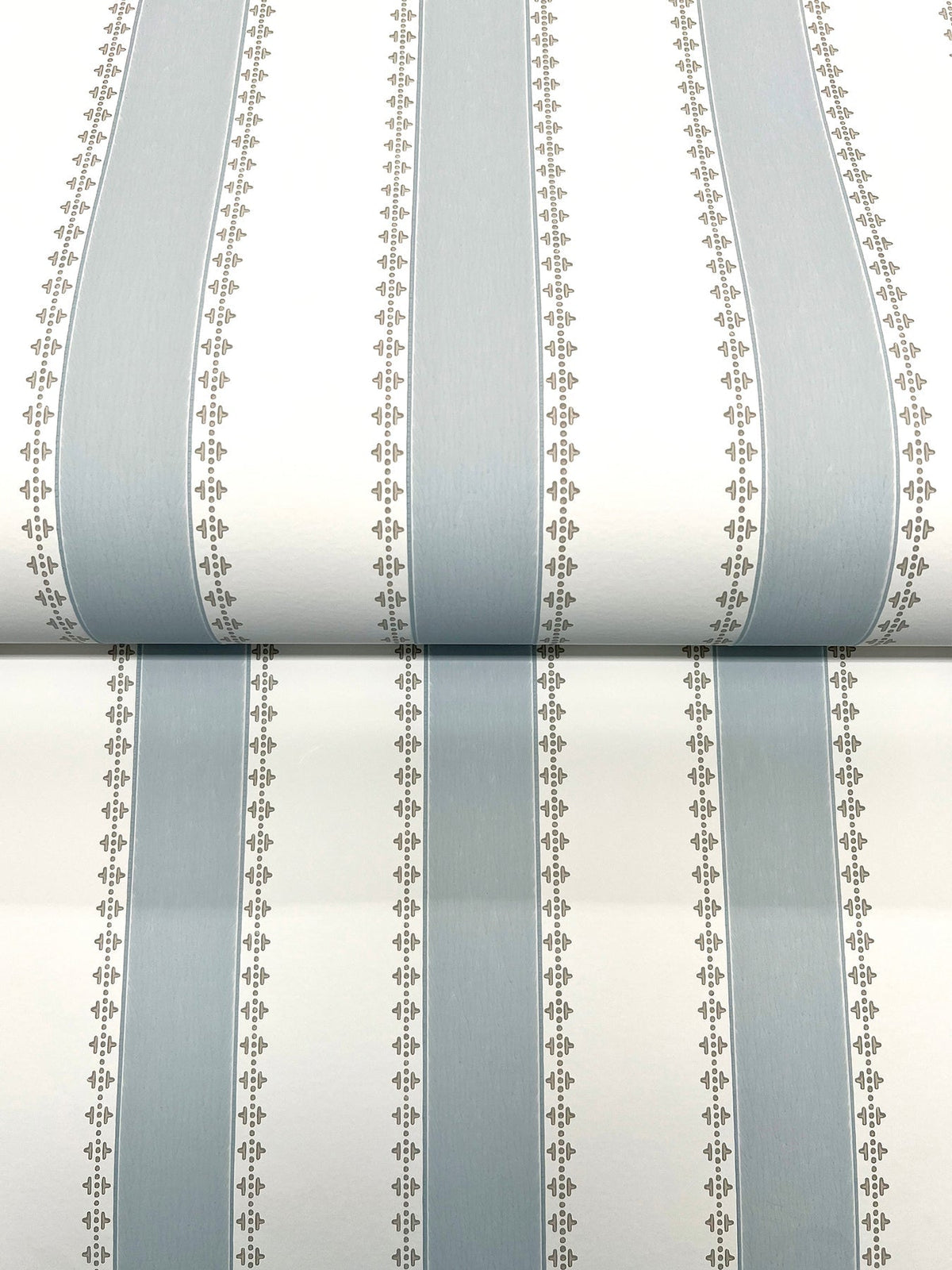 Tilford Awning Stripe Wallpaper by York Wallcoverings features alternating wide light blue and white vertical stripes, separated by narrow beige lines adorned with small leaf patterns. The photo displays a section of the wallpaper rolled out.