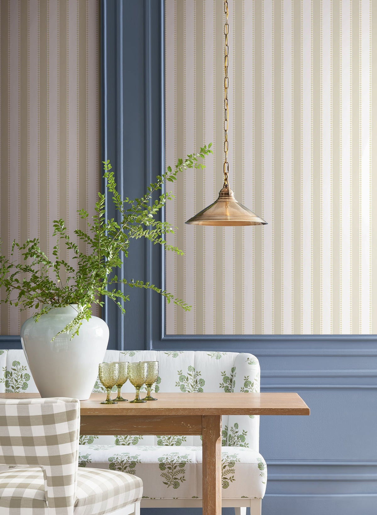 A dining nook featuring a wooden table, a checkered chair, a floral upholstered bench, a white vase filled with green branches, glassware, a brass pendant light, and Tilford Awning Stripe Wallpaper by York Wallcoverings paired with blue wall paneling.