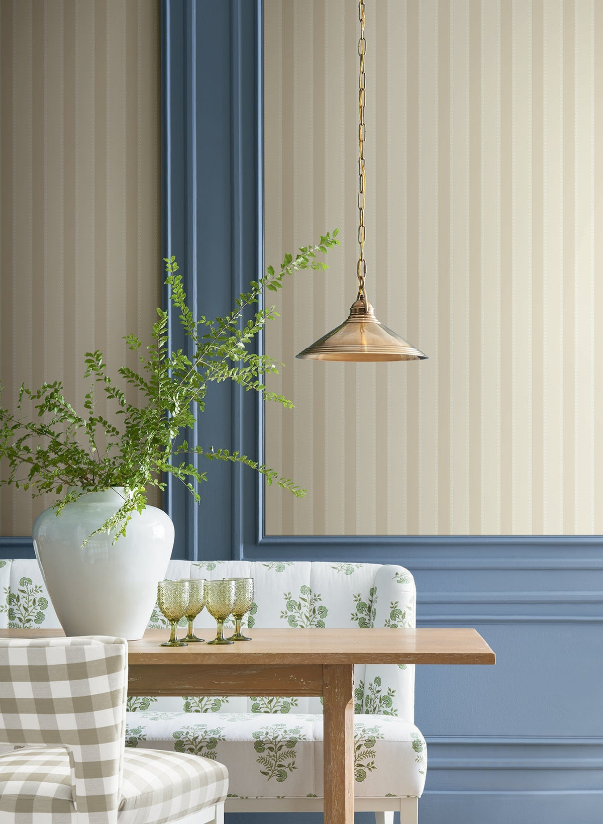 A brass pendant light hangs over a wooden table with a white vase of green branches, three green glasses, and patterned seating, set against walls adorned with York Wallcoverings’ Tilford Awning Stripe Wallpaper featuring beige stripes and blue paneling.