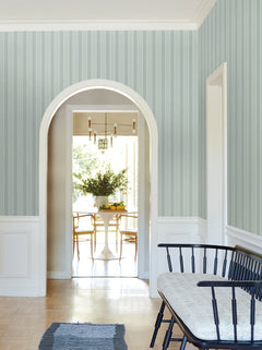 A hallway features Tilford Awning Stripe Wallpaper by York Wallcoverings in blue, a black bench with a cushion, and a blue rug. An arched doorway opens to a dining area furnished with a round white table, wooden chairs, and a vase of greenery.