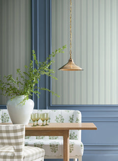 A wooden table with a white vase of green branches, four green glasses, and patterned seating sits below a brass pendant lamp and in front of walls adorned with York Wallcoverings’ Tilford Awning Stripe Wallpaper.