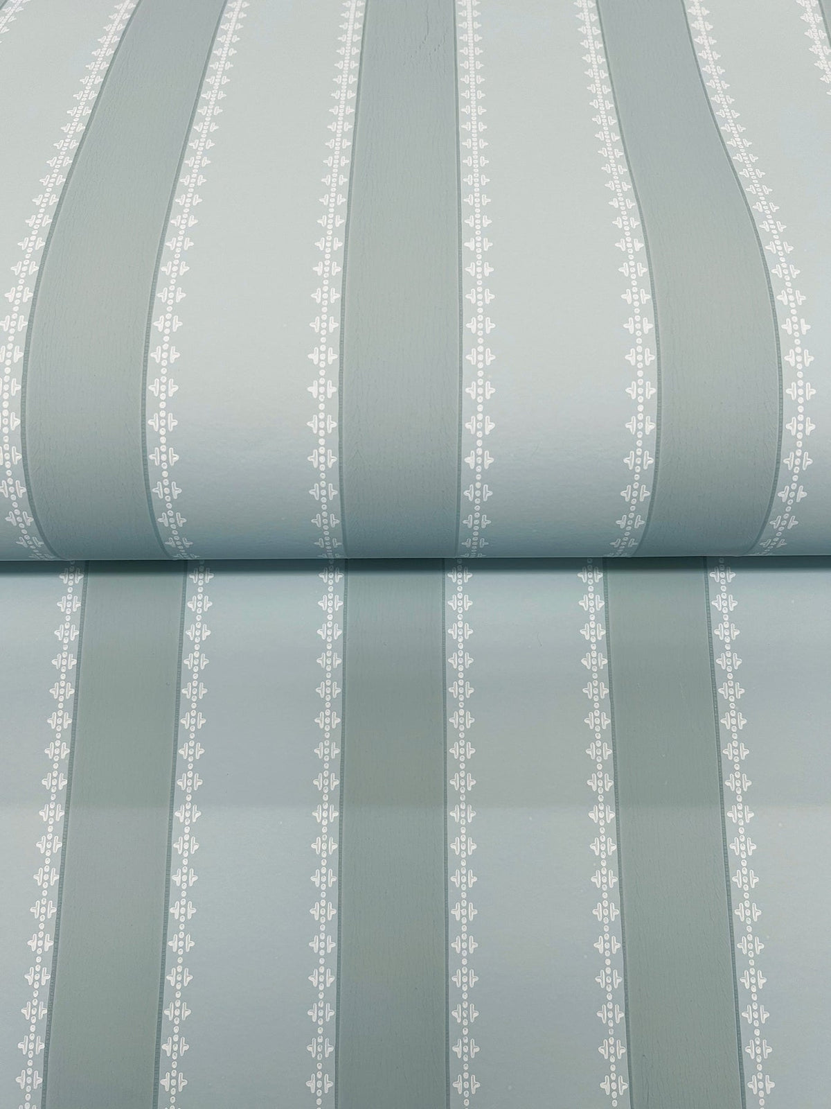 The image shows York Wallcoverings’ Tilford Awning Stripe Wallpaper, featuring light blue with wide, vertical sage-green stripes and thin white lines accented by small leaf patterns on each side. The wallpaper is rolled out with a visible horizontal seam across the center.