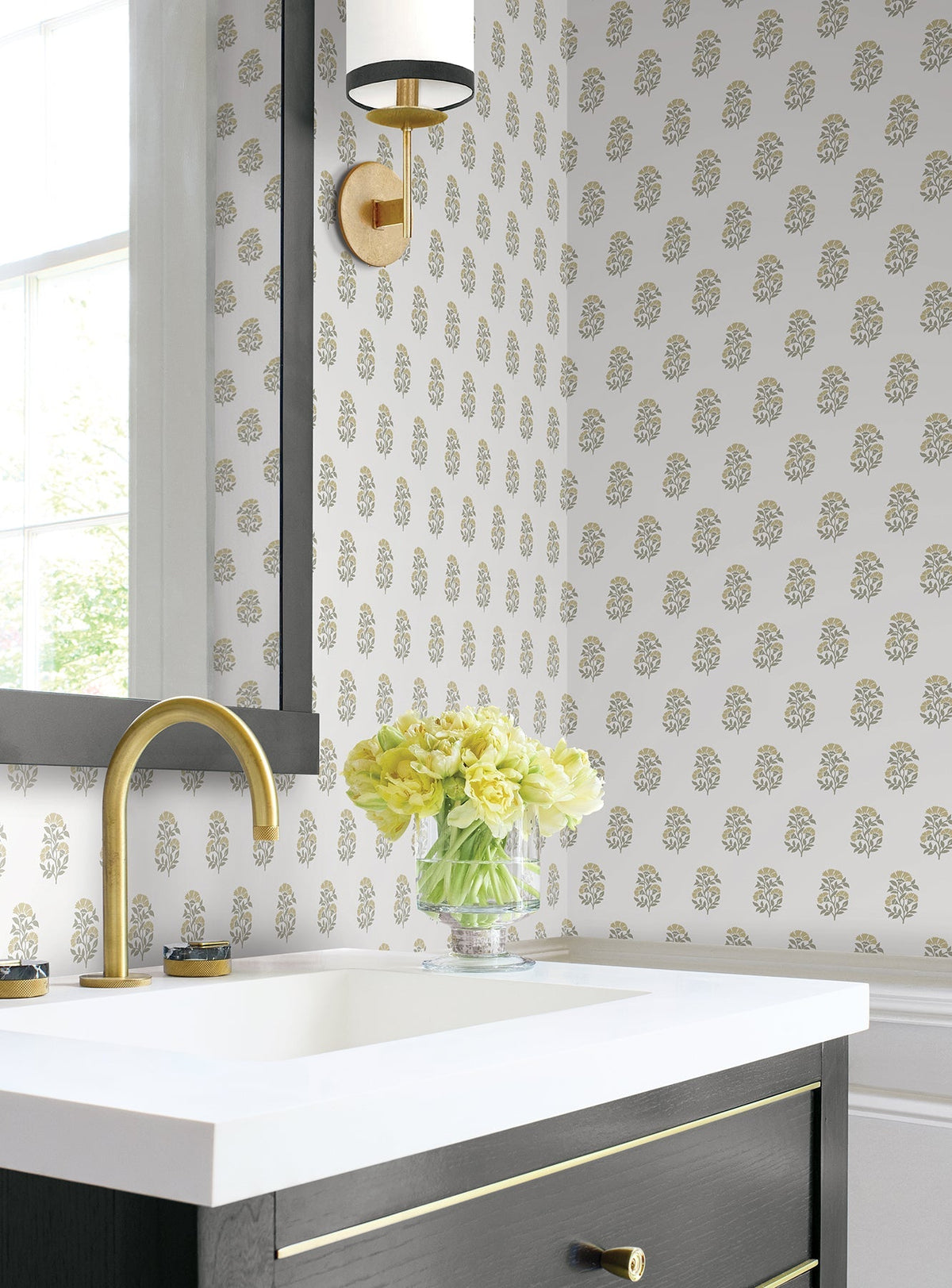 A bathroom featuring York Wallcoverings’ Sylvie Floral Wallpaper, a gold faucet, a glass vase with yellow flowers on the counter, a rectangular mirror, and a gold and black wall sconce beside a window.