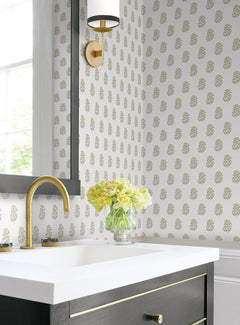A modern bathroom features York Wallcoverings’ Sylvie Floral Wallpaper, gold accents, a black vanity with a white countertop, and a glass vase filled with yellow flowers next to a gold faucet. Sunlight pours in through a nearby window.