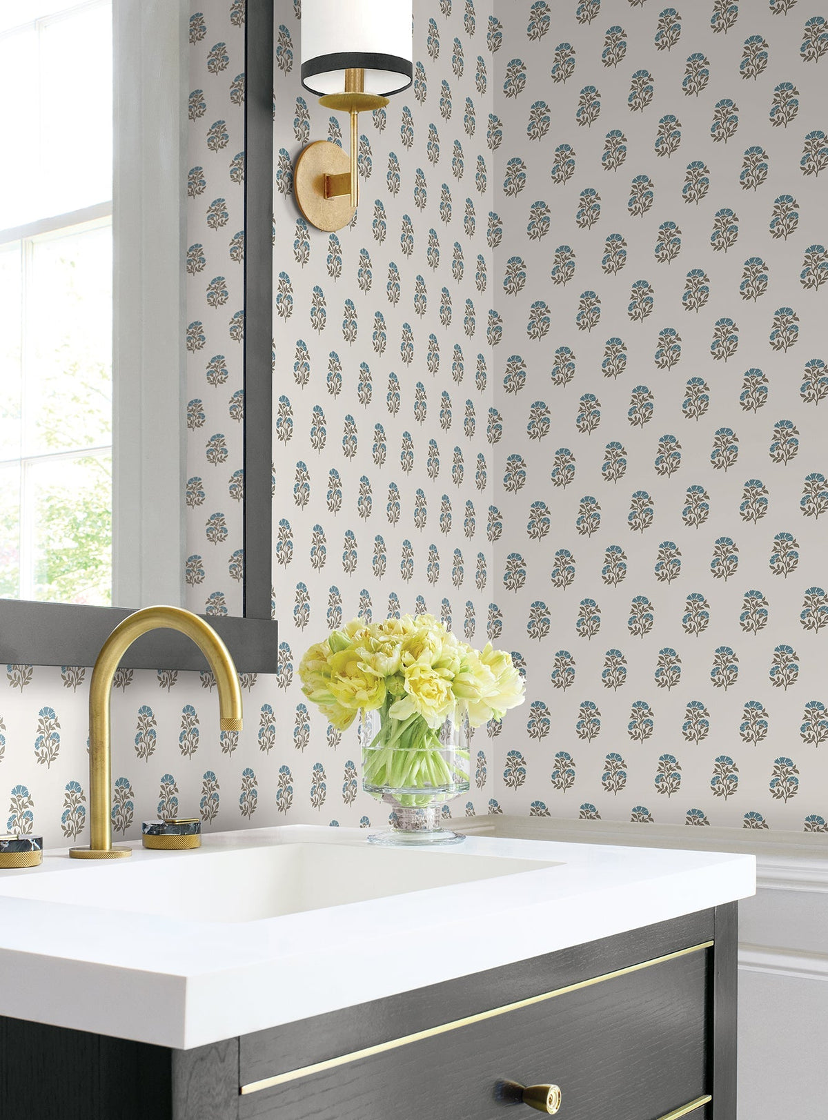 A bathroom featuring Sylvie Floral Wallpaper by York Wallcoverings, a gold faucet, a white sink, a black vanity with a gold drawer pull, a wall sconce, and a vase of yellow flowers beside the window.