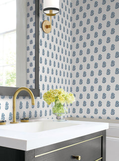 A modern bathroom featuring Sylvie Floral Wallpaper by York Wallcoverings in blue, complemented by a gold faucet and a glass vase of yellow roses on a white countertop beneath a wall-mounted light fixture.