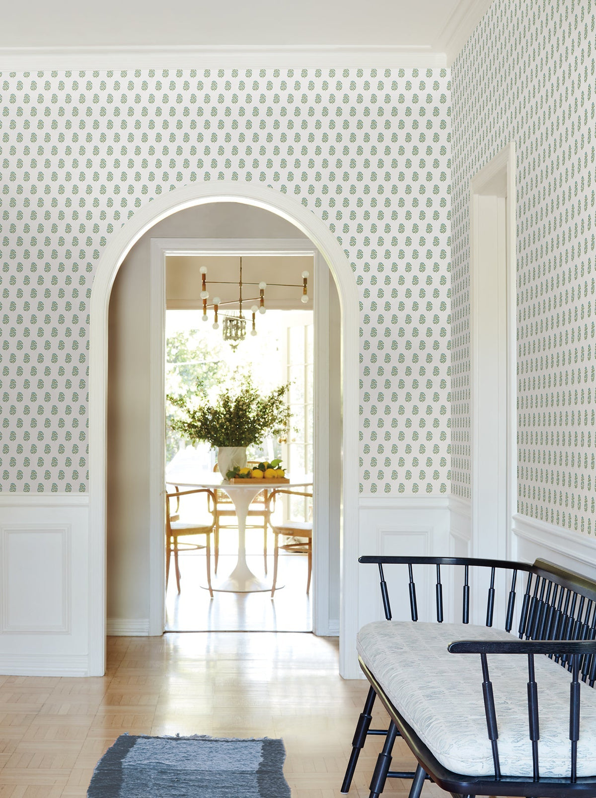 A bright hallway featuring Sylvie Floral Wallpaper by York Wallcoverings, a black bench with a cushion, and an arched doorway leading to a dining room with a round table, chairs, a chandelier, and a large bouquet of flowers.