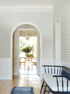A bright hallway featuring Sylvie Floral Wallpaper by York Wallcoverings, a black bench with a cushion, and an arched doorway leading to a dining room with a round table, chairs, a chandelier, and a large bouquet of flowers.