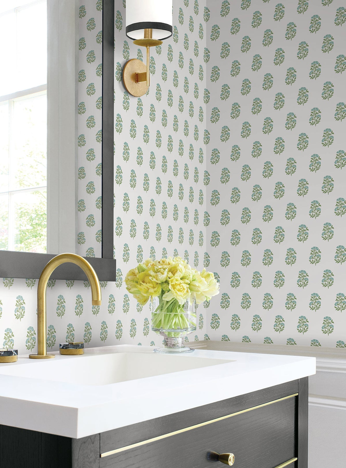 A bathroom featuring Sylvie Floral Wallpaper by York Wallcoverings, a gold faucet, white sink, black vanity, brass sconce, and a glass vase of yellow flowers on the counter near a window.