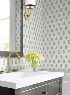 A bathroom featuring Sylvie Floral Wallpaper by York Wallcoverings, a gold faucet, white sink, black vanity, brass sconce, and a glass vase of yellow flowers on the counter near a window.