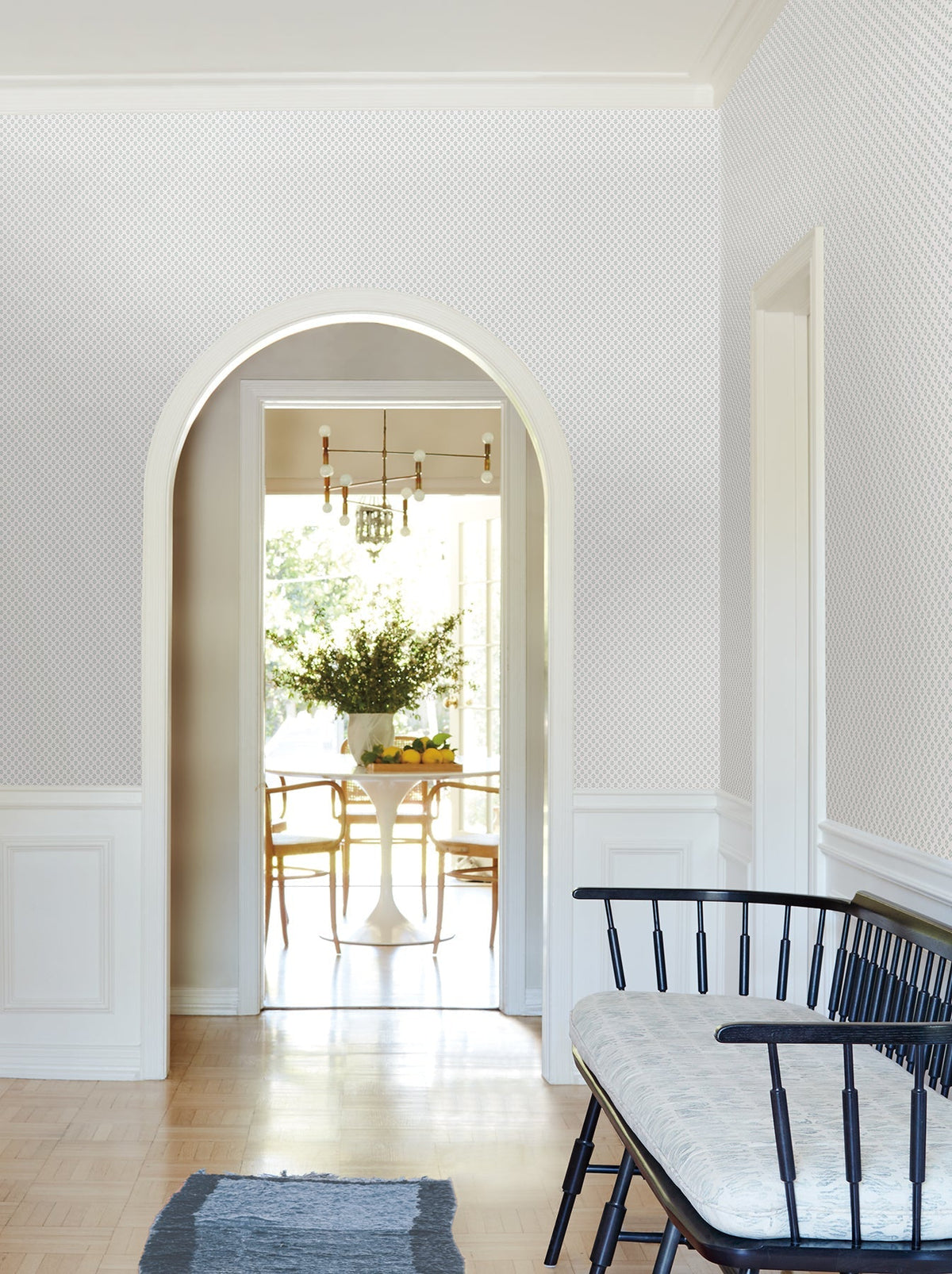 A bright hallway with a black bench leads to a dining area through an arched doorway. The dining room features York Wallcoverings’ Brioni Floral Stamp Wallpaper, a round white table with chairs, a centerpiece of greenery, and large windows that flood the space with natural light.