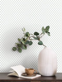 A white vase with green eucalyptus branches sits on a dark wood table next to an open book and a brown ceramic cup. The background features Brioni Floral Stamp Wallpaper by York Wallcoverings, showcasing a delicate green floral pattern.