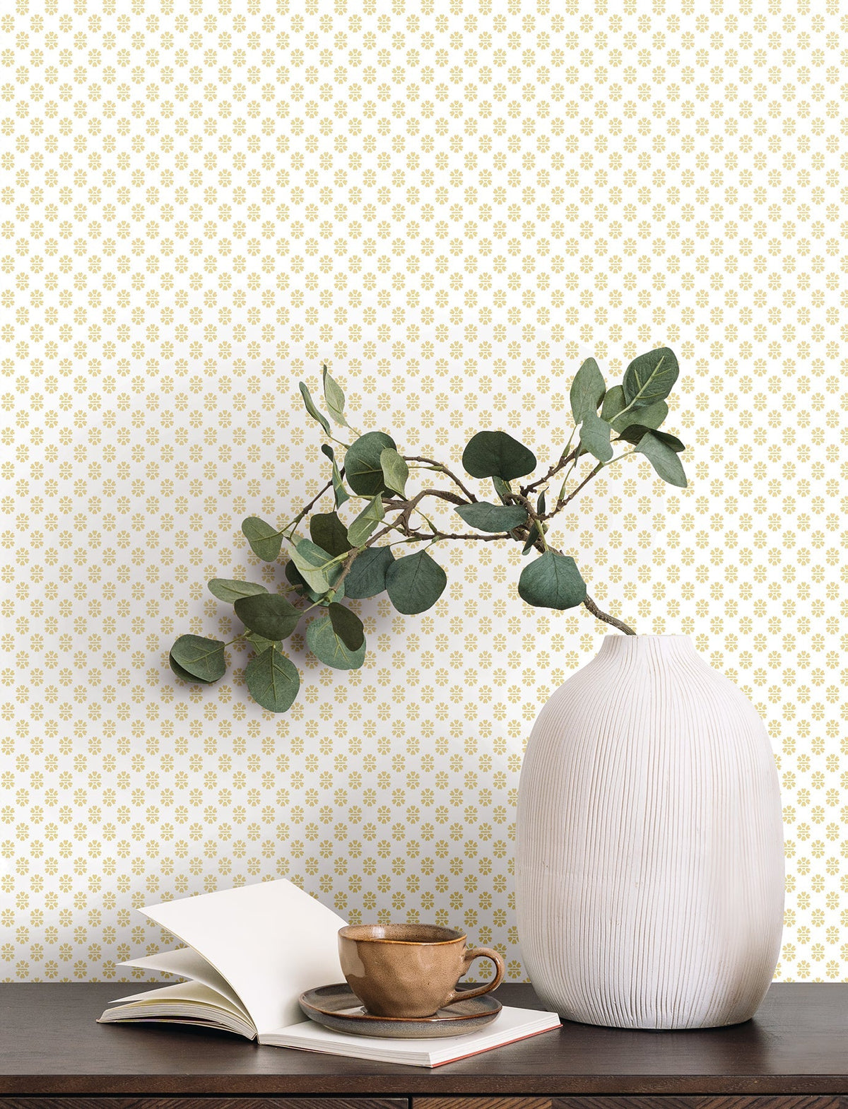 A white ribbed vase with green branches, an open notebook, and a brown cup and saucer sit on a dark wooden table against a wall covered in York Wallcoverings’ Brioni Floral Stamp Wallpaper.