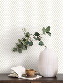 A white vase filled with green eucalyptus branches sits on a dark table beside an open book and a brown cup and saucer, set against a light wall adorned with York Wallcoverings’ Brioni Floral Stamp Wallpaper.