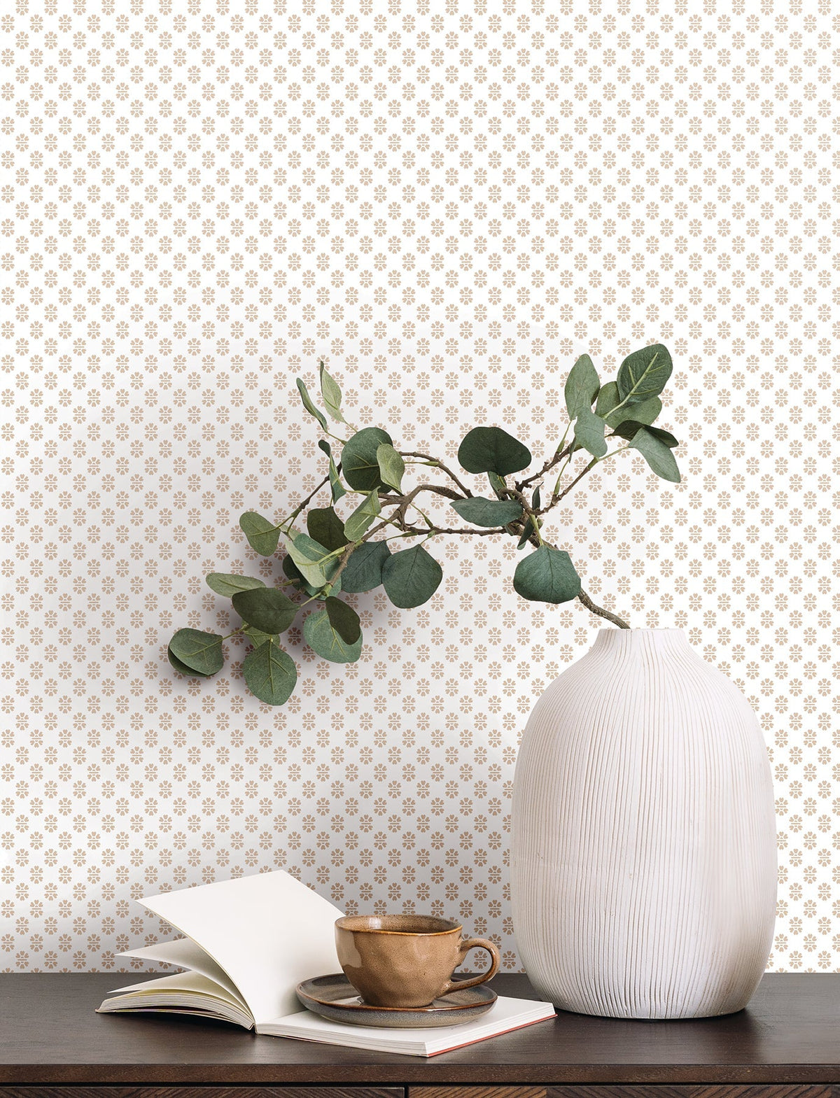A white ribbed vase with green leaves sits on a dark wooden surface next to an open book and a brown cup and saucer. The background features York Wallcoverings’ Brioni Floral Stamp Wallpaper, showcasing a small, repeating gold floral pattern on a white wall.