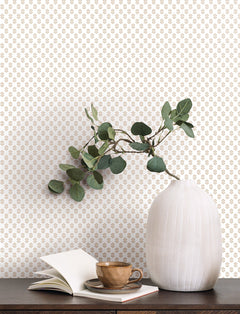 A white ribbed vase with green leaves sits on a dark wooden surface next to an open book and a brown cup and saucer. The background features York Wallcoverings’ Brioni Floral Stamp Wallpaper, showcasing a small, repeating gold floral pattern on a white wall.