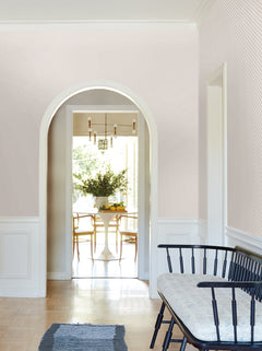 A bright, elegant hallway features a white arched doorway, Brioni Floral Stamp Wallpaper by York Wallcoverings in a light pattern, a black bench with a cushion, and leads into a dining area with a round table, chairs, and a large bouquet of greenery.