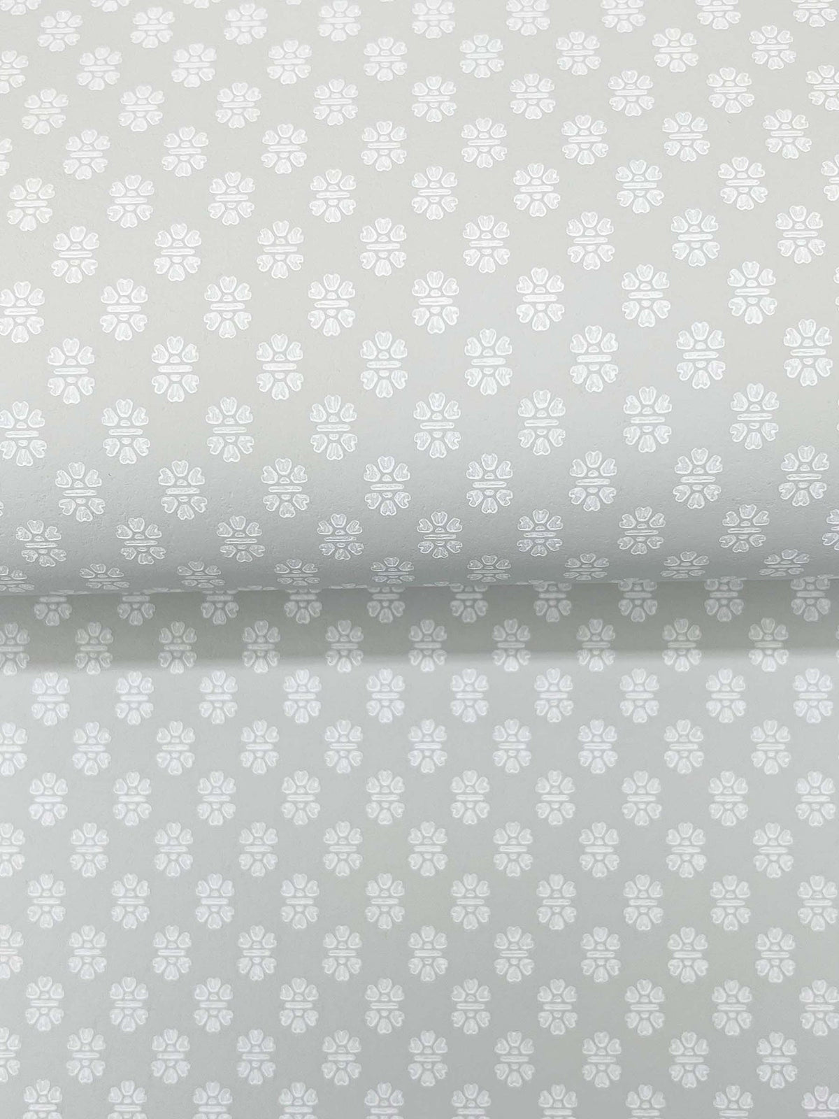 The Brioni Floral Stamp Wallpaper by York Wallcoverings features a light grey backdrop adorned with a subtle, repeating pattern of small white four-petaled flowers, creating a soft, delicate, and uniform appearance across the surface.