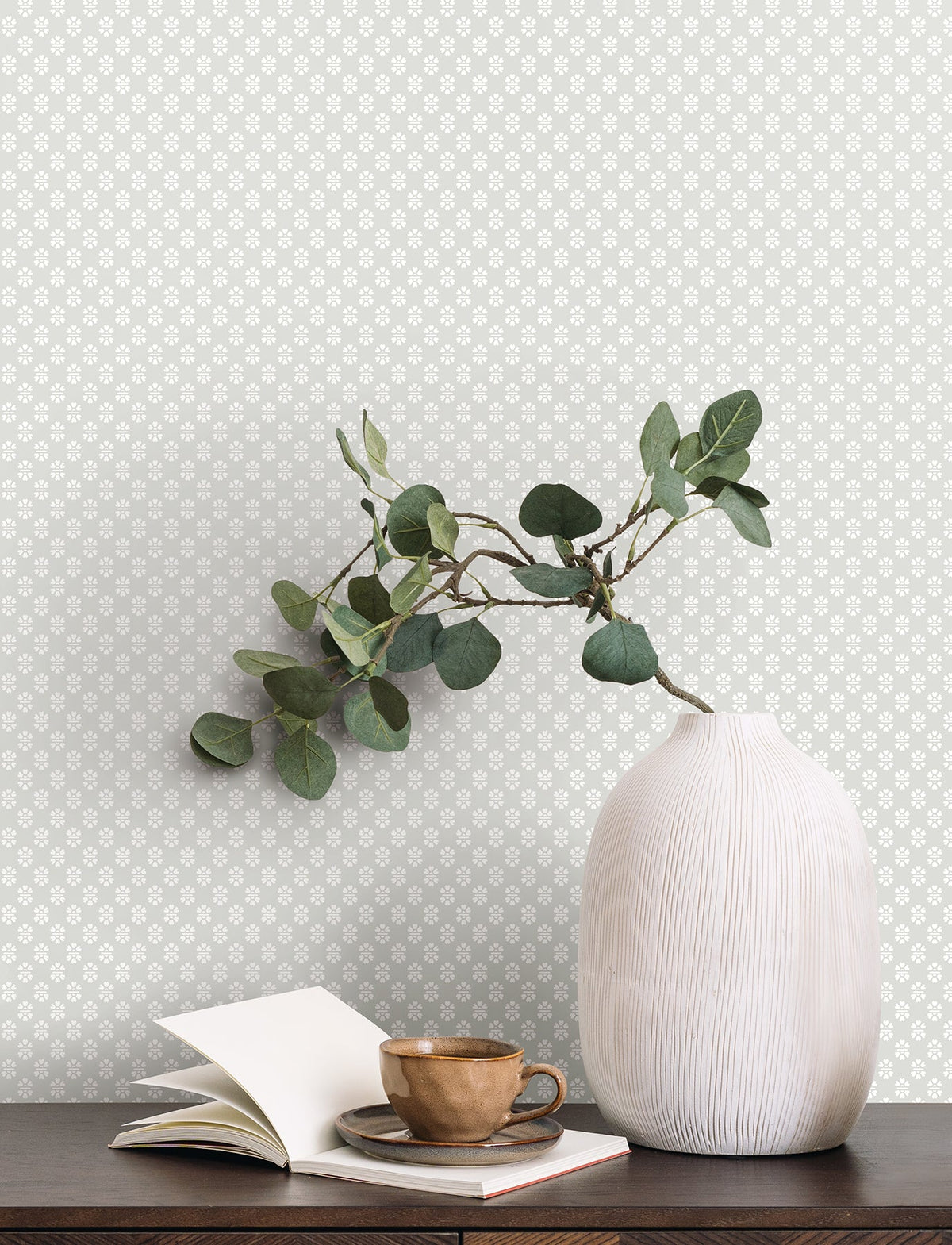 A white ribbed vase with leafy branches sits on a dark table next to a brown teacup, saucer, and an open notebook. The background features York Wallcoverings’ Brioni Floral Stamp Wallpaper in a light color with a small, repeating floral pattern.
