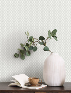 A white ribbed vase with leafy branches sits on a dark table next to a brown teacup, saucer, and an open notebook. The background features York Wallcoverings’ Brioni Floral Stamp Wallpaper in a light color with a small, repeating floral pattern.