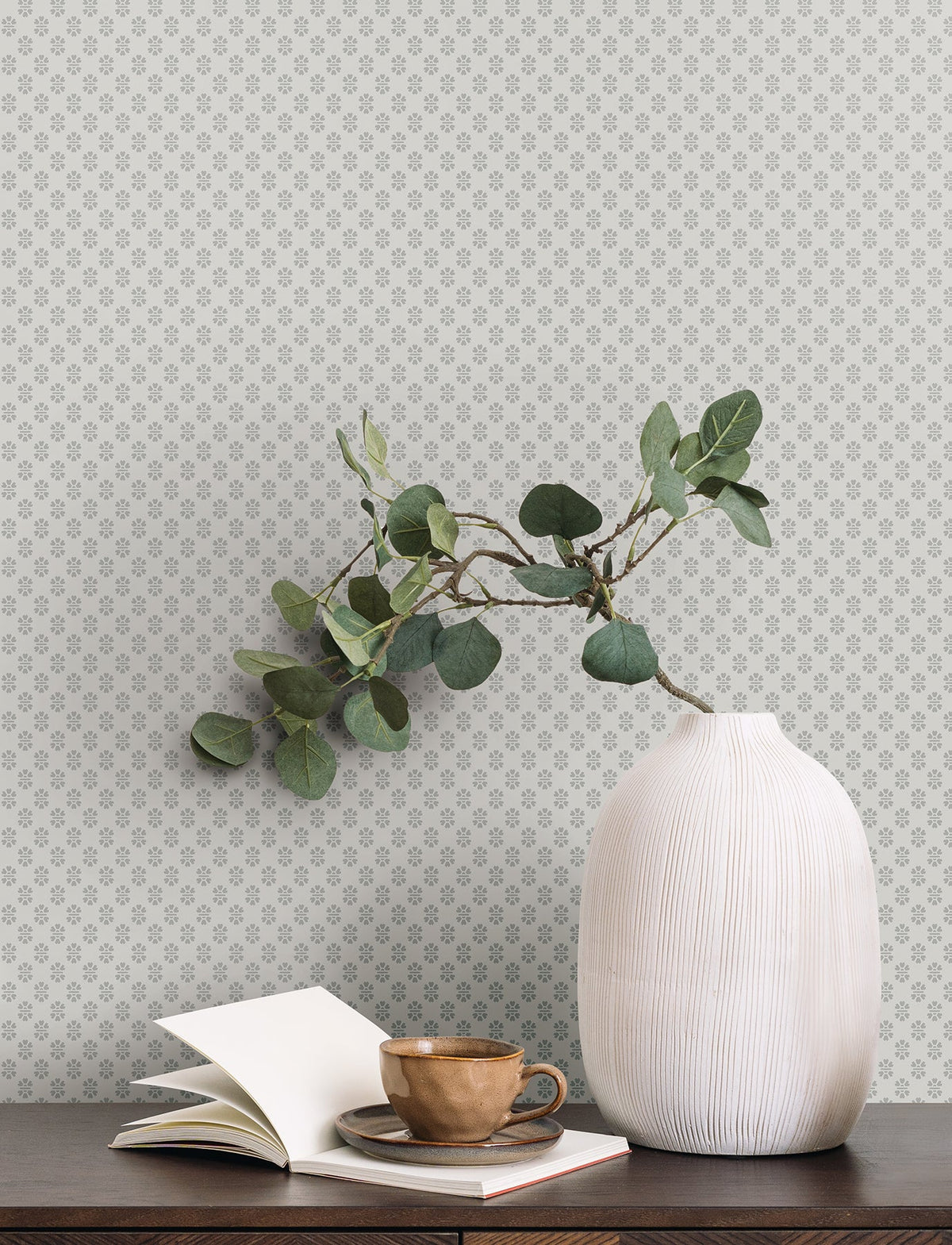 A white ribbed vase with green leafy branches sits on a dark wood table beside an open book, a closed book, and a brown ceramic cup, set against York Wallcoverings’ Brioni Floral Stamp Wallpaper featuring a light background with a small, repeating floral pattern.