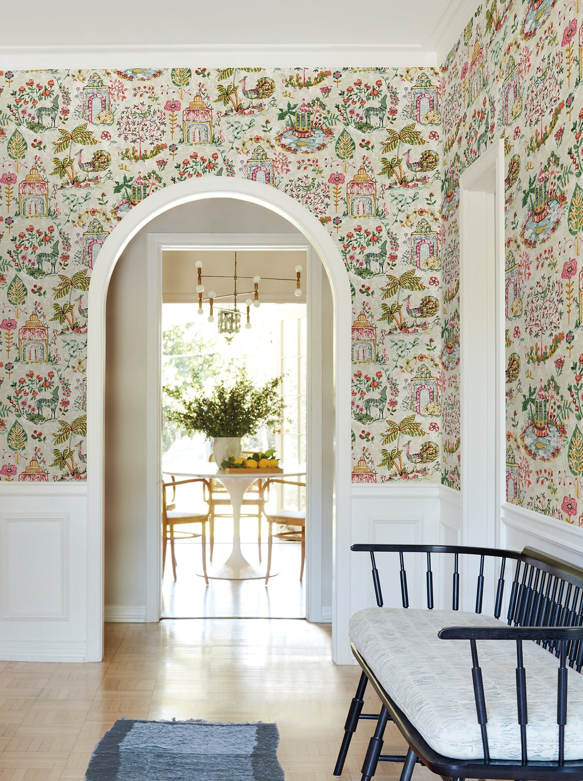 A hallway adorned with Parc Garden Dreams Wallpaper by York Wallcoverings, featuring white trim, a black bench with a cushion, and an arched doorway that opens into a bright dining area with a round table, chairs, and a vase of flowers.