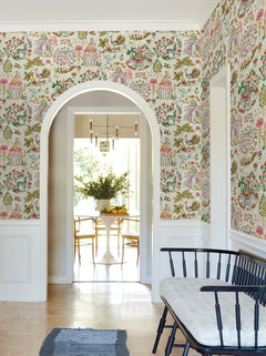 A hallway adorned with York Wallcoverings’ Parc Garden Dreams Wallpaper—a colorful, floral design—features white trim and an arched doorway that opens to a dining room furnished with a round white table and wooden chairs. Along the hallway wall sits a black bench topped with a cushion.
