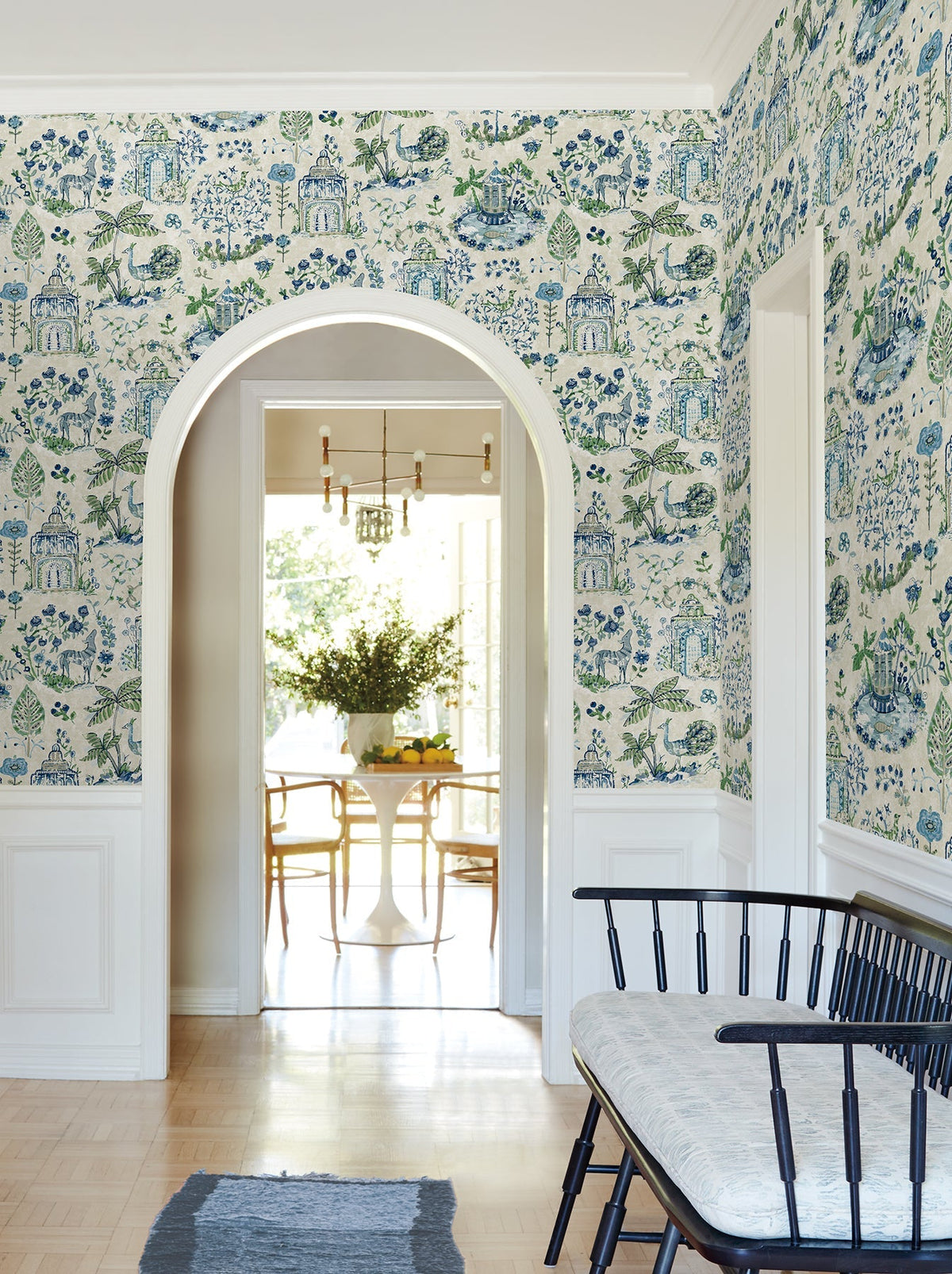 A hallway featuring Parc Garden Dreams Wallpaper by York Wallcoverings in blue and green patterns, a black bench with a light cushion, white trim, and an arched doorway that opens to a bright dining area with a round table and chairs.