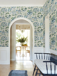A hallway adorned with York Wallcoverings’ Parc Garden Dreams Wallpaper, featuring blue and white patterns, leads through an arched doorway to a bright dining area with a round white table, chairs, and a vase of flowers. A cushioned black bench sits against the wall.