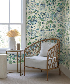 A wicker chair with a white cushion sits by a window next to a small round table adorned with vases and dried flowers. The walls are covered in York Wallcoverings’ Parc Garden Dreams Wallpaper, featuring a blue and green botanical and gazebo pattern. The room has a beige carpeted floor.