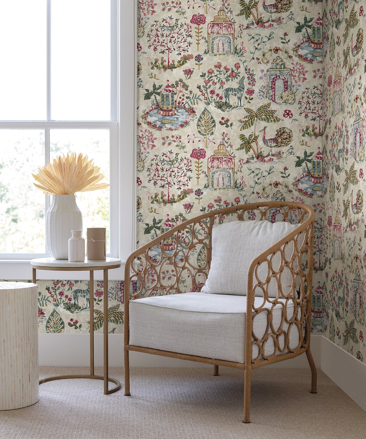A cozy corner featuring a wicker chair with a cushion, a small round table adorned with vases and dried grasses, and York Wallcoverings’ Parc Garden Dreams Wallpaper in a floral pattern near a large window filled with natural light.