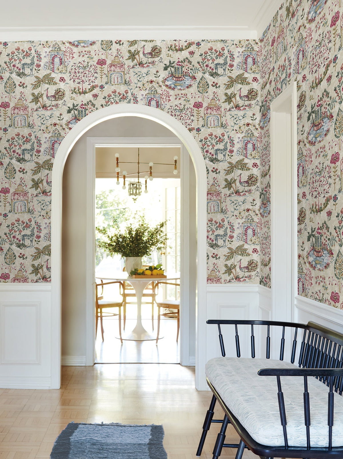 A hallway adorned with York Wallcoverings’ Parc Garden Dreams Wallpaper, featuring an archway that opens to a bright dining area with a round table and chairs, a black bench topped with a cushion, and a rug resting on light wood floors.