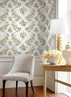 A stylish room featuring Genevieve Floral Trail Wallpaper by York Wallcoverings, a patterned white chair, a wooden table with a gold lamp, a white vase holding a bouquet of yellow roses, all set against elegant white wainscoting and hardwood floors.