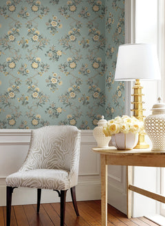A chair with a light pattern sits beside a wooden table topped with a lamp, a jar, and a vase of yellow roses. The room features Genevieve Floral Trail Wallpaper by York Wallcoverings in blue, large windows that flood the space with light, wood floors, and classic white wainscoting for an elegant look.