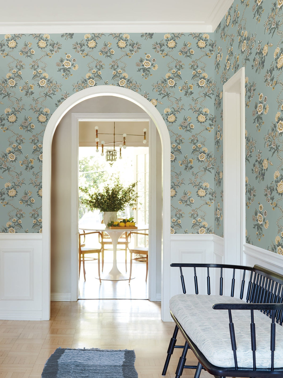 A hallway adorned with York Wallcoverings’ Genevieve Floral Trail Wallpaper in blue and white trim features an arched doorway leading to a bright dining area with a round table, chairs, a vase of flowers, and modern light fixtures. A bench sits against the wall in the foreground.