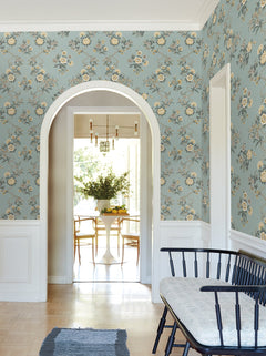 A bright hallway features York Wallcoverings’ Genevieve Floral Trail Wallpaper in blue, an arched doorway, a black bench with a cushion, a rug, and offers a view into a dining area with a white table, wooden chairs, and a vase of greenery.