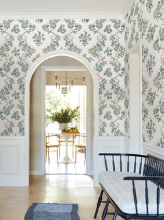 A hallway with Genevieve Floral Trail Wallpaper by York Wallcoverings, a black bench with a cushion, a blue rug, and an arched doorway leading to a sunlit dining area with a round table and chairs.