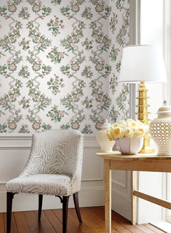 A white chair with a subtle zebra pattern sits next to a wooden table topped with a gold lamp, white jar, and a bouquet of yellow roses. The bright room is decorated with York Wallcoverings’ Genevieve Floral Trail Wallpaper and white paneling.