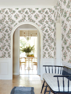 A hallway adorned with Genevieve Floral Trail Wallpaper by York Wallcoverings features a black bench with a cushion, while an arched doorway reveals a sunlit dining area furnished with a round white table, wooden chairs, and a vase of flowers.