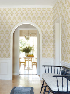 A hallway adorned with York Wallcoverings’ Souci Marigolds Wallpaper in yellow and accented by white trim leads to a bright dining room featuring a round table, chairs, and a vase of greenery. Along the hallway wall, a black bench with a cushion completes the scene.