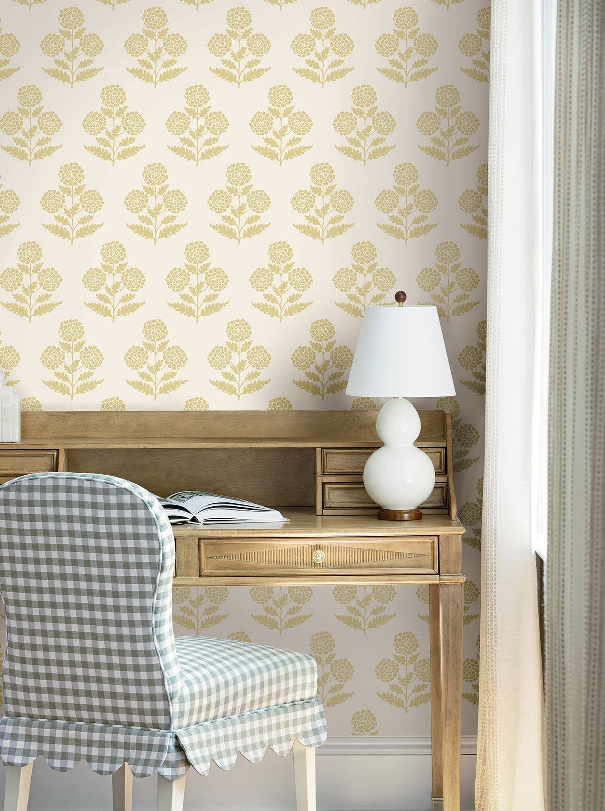 A wooden desk with an open book and a white lamp sits against a wall adorned with York Wallcoverings’ Souci Marigolds Wallpaper. At the desk, a chair upholstered in blue and white gingham fabric is positioned, while light curtains frame a nearby window.