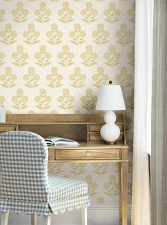 A wooden desk with an open book and a white lamp sits against a wall adorned with York Wallcoverings’ Souci Marigolds Wallpaper. At the desk, a chair upholstered in blue and white gingham fabric is positioned, while light curtains frame a nearby window.