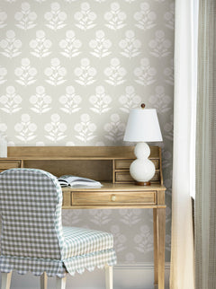 A wooden desk with an open book and white lamp sits against York Wallcoverings’ Souci Marigolds wallpaper. A chair with blue gingham fabric is in front of the desk, and light curtains hang to the right.