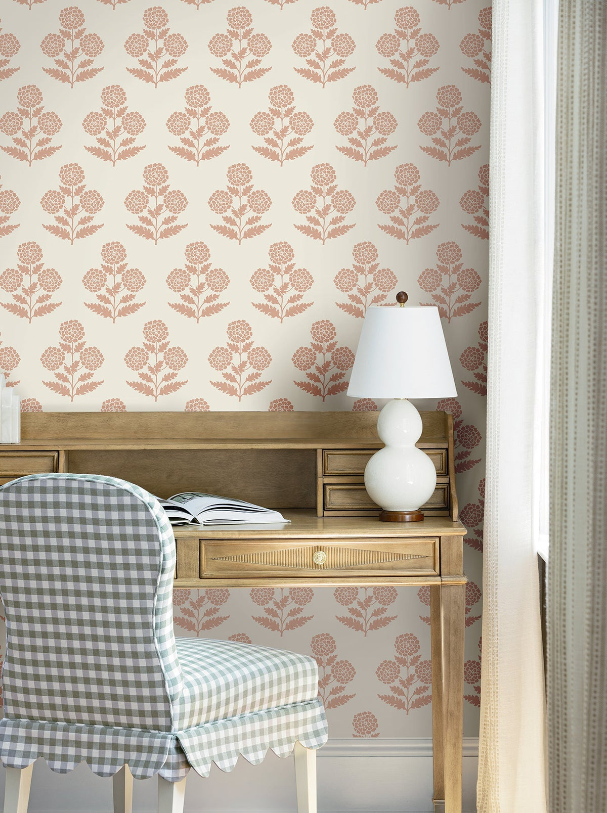 A wooden desk with an open book, a white lamp, and a checkered chair sits in front of a window. The wall behind is adorned with York Wallcoverings’ Souci Marigolds Wallpaper, featuring a pink floral pattern on a cream background. Light curtains frame the scene.