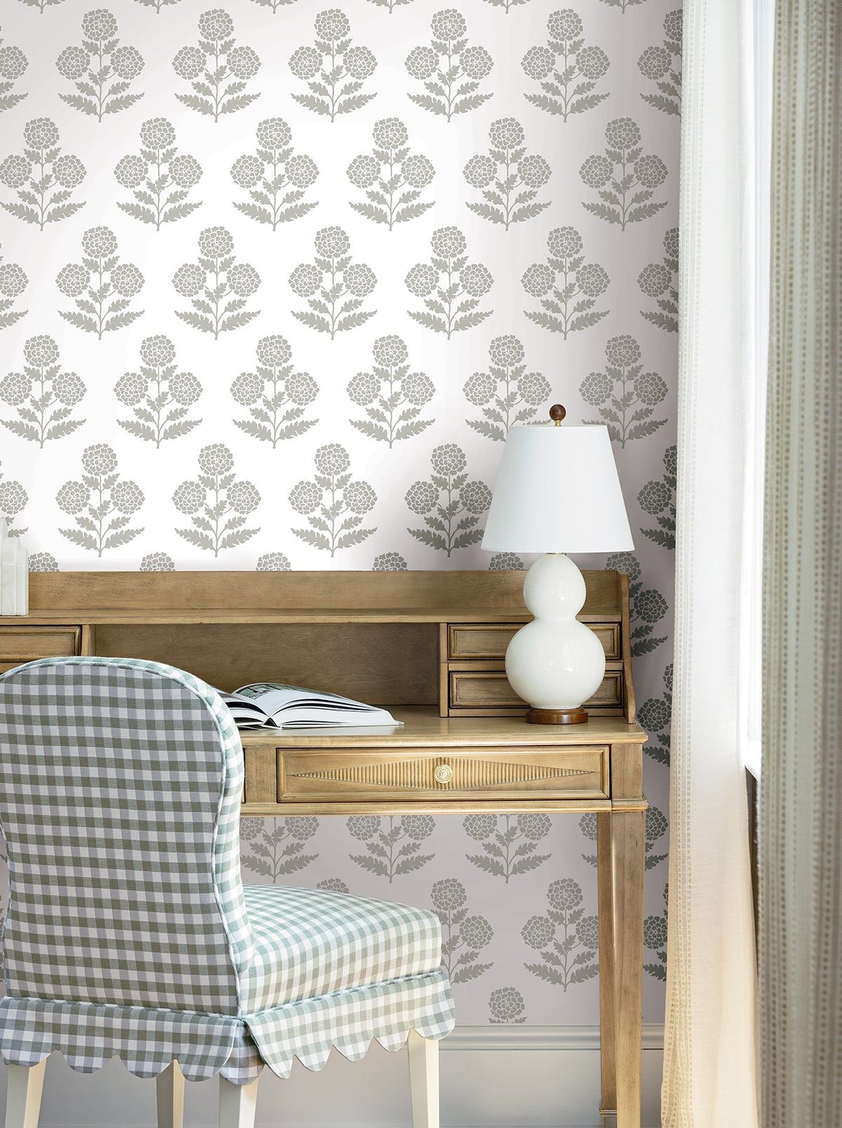 A wooden desk with an open book, white lamp, and checkered chair sits by a window with light curtains. The wall features the Souci Marigolds Wallpaper by York Wallcoverings in muted gray tones.