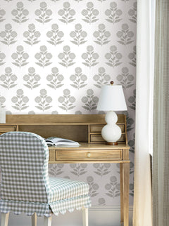 A wooden desk with an open book, a white lamp, and a chair with a blue checkered cover sits in front of a wall adorned with York Wallcoverings’ Souci Marigolds Wallpaper. Light curtains frame the scene.
