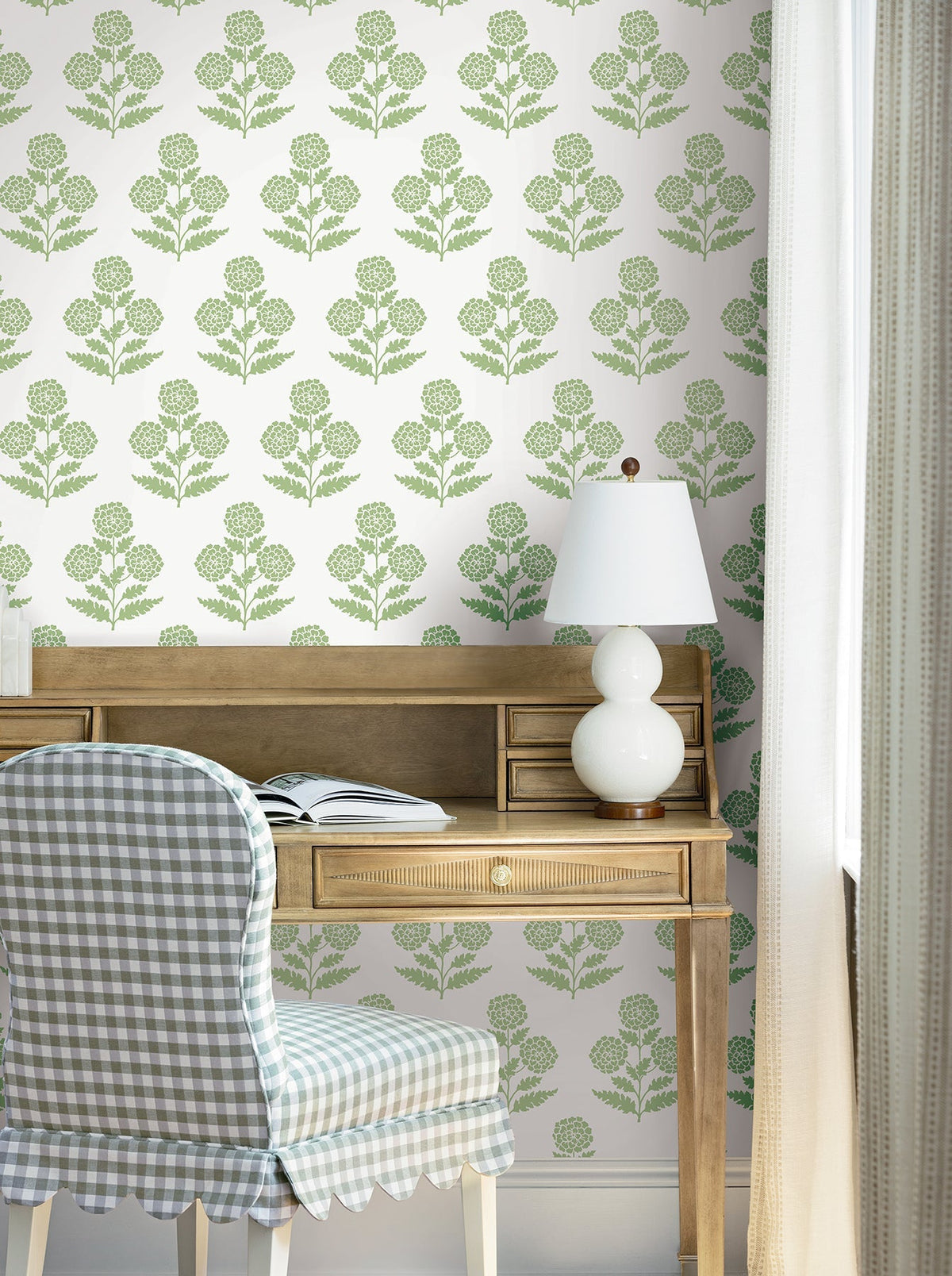 A wooden desk with an open book, a white lamp, and a chair with a blue gingham cover sits in front of a wall adorned with York Wallcoverings’ Souci Marigolds Wallpaper, next to a white curtain.