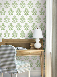 A wooden desk with an open book, a white lamp, and a checked chair sits in front of York Wallcoverings’ Souci Marigolds Wallpaper and airy sheer curtains, creating a cozy, light-filled workspace.