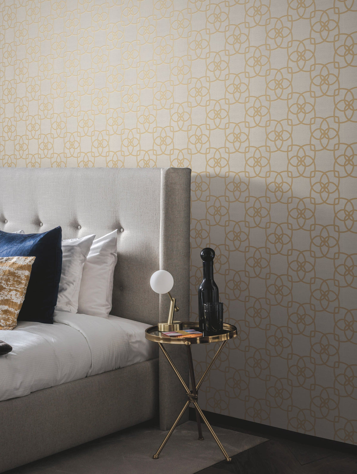 A modern bedroom featuring a beige tufted headboard, gold and blue pillows, and a round gold side table adorned with a lamp, books, and a dark bottle, set against York Wallcoverings’ Serendipity Wallpaper with elegant gold geometric patterns.