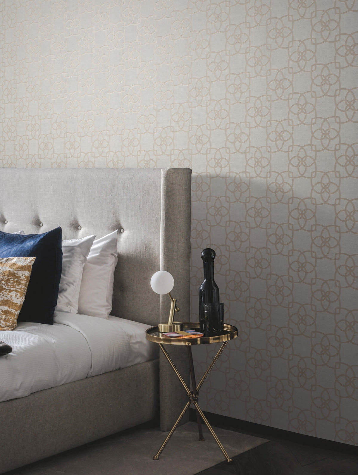 A modern bedroom featuring a gray upholstered bed adorned with blue and gold pillows, a round gold side table displaying a lamp, black bottle, and book, set against York Wallcoverings’ Serendipity Wallpaper in beige.